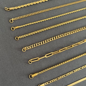 K&ouml;nnte beinhalten: Eine Auswahl goldfarbener Halsketten mit verschiedenen Kettenarten, darunter Seil-, Panzer- und B&uuml;roklammer-Designs. Jede Halskette hat einen Verschluss. Die Ketten sind auf einer grauen Oberfl&auml;che angeordnet.
