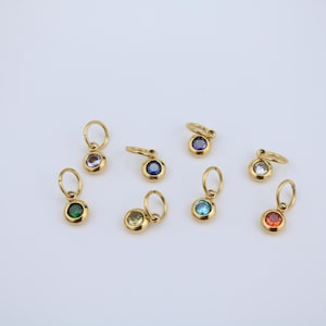 May include: A collection of gold-colored pendants with various colored gemstones. Each pendant features a small, round gemstone in colors like green, blue, purple, and red, set in a gold bezel. The pendants have a small loop for attaching to a chain.