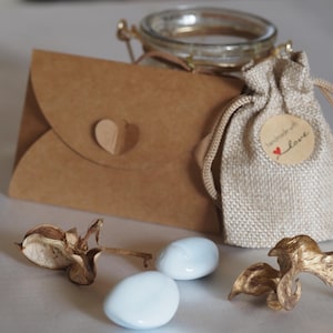 K&ouml;nnte beinhalten: Eine rustikale Anordnung mit einem kleinen Glas, einem Jutebeutel mit einem "handmade with love"-Etikett und einem braunen Papierumschlag mit Herz. Zwei hellblaue, ovale S&uuml;&szlig;igkeiten und getrocknete Blumenelemente.