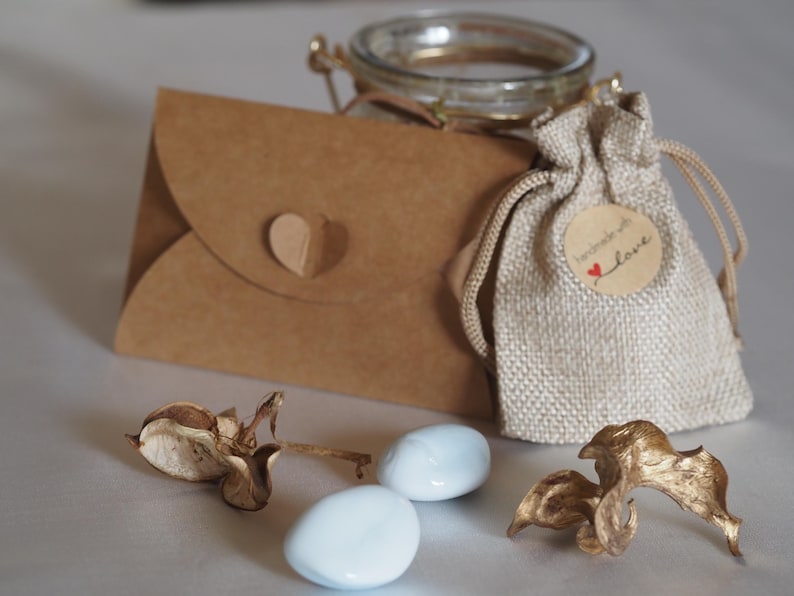 K&ouml;nnte beinhalten: Eine rustikale Anordnung mit einem kleinen Glas, einem Jutebeutel mit einem "handmade with love"-Etikett und einem braunen Papierumschlag mit Herz. Zwei hellblaue, ovale S&uuml;&szlig;igkeiten und getrocknete Blumenelemente.