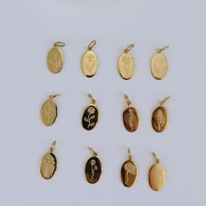 May include: A collection of twelve gold-colored oval pendants, each engraved with a different floral design. The pendants are arranged on a white surface, showcasing the variety of botanical illustrations. The pendants have a small loop at the top for attaching to a chain.