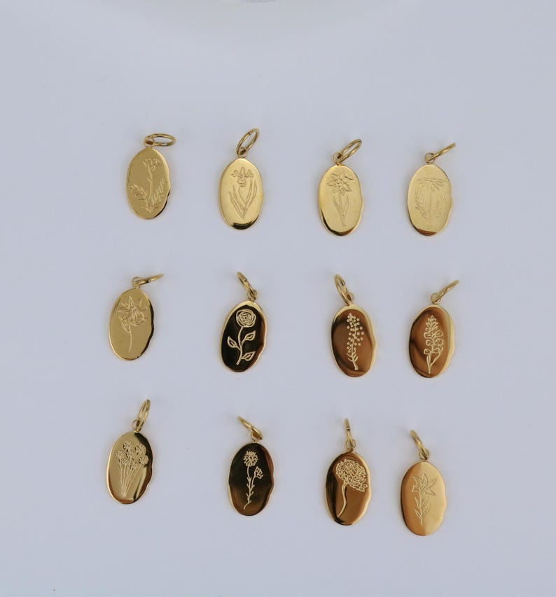 May include: A collection of twelve gold-colored oval pendants, each engraved with a different floral design. The pendants are arranged on a white surface, showcasing the variety of botanical illustrations. The pendants have a small loop at the top for attaching to a chain.