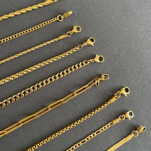 May include: An assortment of gold-colored chain necklaces with various link styles, including rope, curb, and paperclip designs. Each necklace features a lobster clasp closure. The chains are displayed on a gray background.