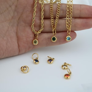 May include: Gold-colored necklaces with gemstone pendants in various colors, including green and blue. Several pairs of gold earrings with gemstone accents are also visible. The jewelry is displayed on a white surface.