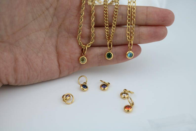 May include: Gold-colored necklaces with gemstone pendants in various colors, including green and blue. Several pairs of gold earrings with gemstone accents are also visible. The jewelry is displayed on a white surface.