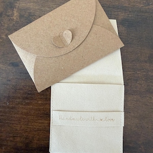 K&ouml;nnte beinhalten: Ein hellbeige Kartenhalter mit passendem Umschlag. Der Umschlag hat einen herzf&ouml;rmigen Verschluss. Auf dem Kartenhalter steht "Handmade with love".