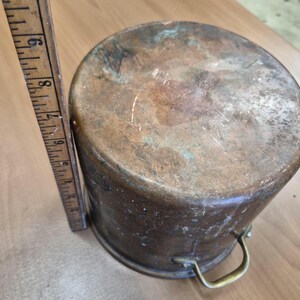 Può includere: Una pentola cilindrica in rame con una superficie usurata e ossidata, dotata di un unico manico in ottone. Un righello in legno è posizionato verticalmente accanto alla pentola, indicando un'altezza di circa 23 centimetri dalla base al bordo.