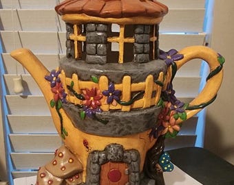 May include: A ceramic teapot shaped like a whimsical house with a brown roof, stone walls, and a yellow base. The teapot is decorated with flowers, vines, and a small door.