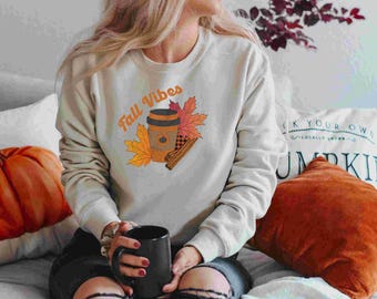 Może przedstawiać: Jasnoszara bluza z napisem "Fall Vibes" i grafiką filiżanki kawy, jesiennych liści i lasek cynamonu. W dłoniach trzymany jest czarny kubek. Pomarańczowa dynia i poduszka znajdują się w tle.