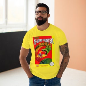 Może przedstawiać: Żółta koszulka z grafiką gry NES 'Galaxy Burai Fighter'. Grafika przedstawia smoka i postać walczącą.