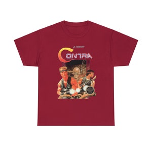 May include: A red t-shirt with a vintage video game graphic of the game "Contra" featuring two soldiers and an alien. The text "KONAMI" is printed above the graphic.
