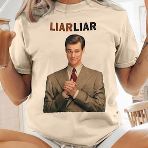 Può includere: T-shirt color crema con la scritta "LIARLIAR" sopra la grafica di un uomo in giacca e cravatta. L'uomo sorride con le mani giunte. La maglietta è di stile casual.