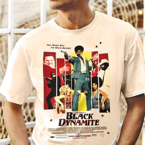 Może przedstawiać: Kremowa koszulka z projektem plakatu filmowego dla "Black Dynamite". Plakat przedstawia mężczyznę w niebieskim garniturze z pistoletem, otoczonego przez inne postacie. Tekst zawiera "They Messed With The Wrong Brother" i "Coming Soon to Theatres."
