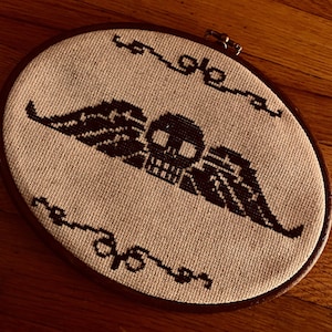 Puede incluir: Un bordado de punto de cruz en blanco y negro de una calavera con alas. El bordado está enmarcado en un aro de madera marrón.
