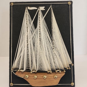 Peut inclure: Une œuvre d'art en fil faite à la main représentant un voilier. Le navire présente des voiles en fil blanc et une coque en bois, sur un fond en feutre noir. Des épingles de couleur laiton soulignent le navire et encadrent l'œuvre, créant un effet texturé et en trois dimensions.