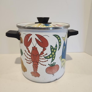 May include: A white enamel cooking pot with a lid and black handles. The pot is decorated with colorful illustrations of a lobster, fish, vegetables, and other food items. The lid has a black knob.