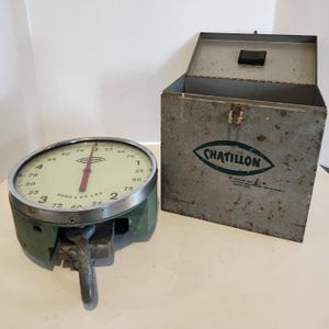 Könnte beinhalten: Eine antike Chatillon-Waage mit grünem Sockel und rundem Zifferblatt. Das Zifferblatt hat schwarze Zahlen und einen roten Zeiger, mit einem Messbereich von 3000 x 25 lbs. Eine passende Metallbox mit dem Chatillon-Logo befindet sich neben der Waage.
