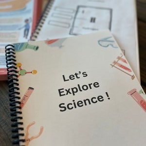 Op de afbeelding: Een wit spiraalnotitieblok met de tekst "Let's Explore Science!" op de omslag. De omslag heeft een lichtroze achtergrond met illustraties van wetenschappelijke apparatuur, waaronder bekers, reageerbuizen en een molecuul.