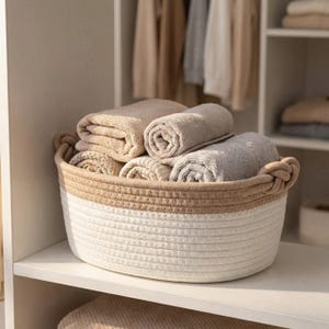 Puede incluir: Una cesta tejida de dos tonos llena de toallas beige y grises enrolladas y dobladas. La cesta tiene una base blanca y una sección superior marrón con asas de cuerda. Las toallas están cuidadosamente dispuestas en el interior, lo que sugiere una solución de almacenamiento.