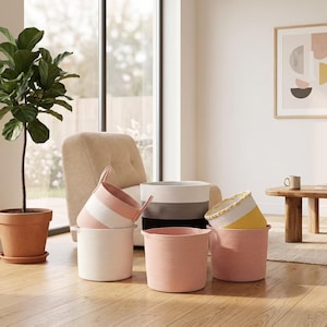 May include: A collection of six cylindrical storage baskets made from coiled rope. The baskets feature various solid and striped color patterns in white, soft pink, dark gray, and yellow. Each basket has a simple, open-top design.