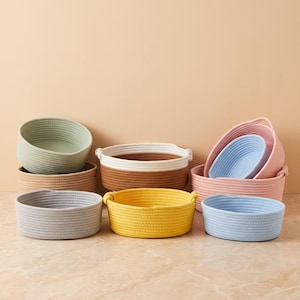 May include: A collection of round storage baskets made from coiled rope in various colors including sage green, tan, brown, white, gray, yellow, light blue, and pink. The baskets are arranged in a group, featuring different sizes and depths.
