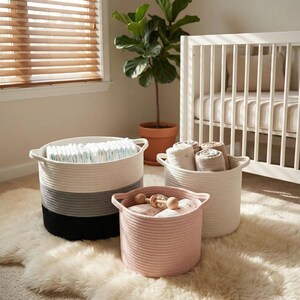 Könnte beinhalten: Drei geflochtene Seilkörbe in einem Kinderzimmer. Der größte Korb ist weiß, grau und schwarz und mit Windeln gefüllt. Ein weißer Korb enthält gerollte Decken und ein rosa Korb Babyartikel.