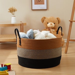 May include: A round, multi-colored storage basket with black handles. The basket is made of woven rope in three horizontal bands: black at the bottom, a tan and black pattern in the middle, and a solid tan at the top. The basket is filled with soft toys.