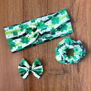 May include: A St. Patrick's Day themed hair accessory set. The set includes a headband, a scrunchie, and a bow. Each item features a white background with green shamrocks, gold dots, and black and green stripes.