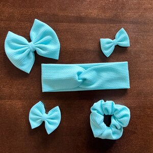 May include: A set of light blue hair accessories, including two bow hair clips, a headband, and a scrunchie. The items are made of a textured fabric and are arranged on a brown surface. The bows vary in size, offering options for different hairstyles.