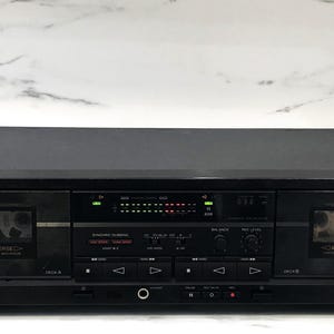 May include: A black Sony stereo cassette deck featuring dual tape compartments labeled Deck A and Deck B with auto-reverse functionality. The front panel includes a central digital display, various control buttons, a headphone jack, and volume knobs for recording levels and balance adjustments.