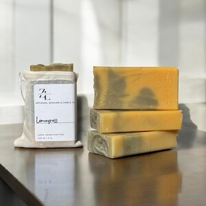May include: Four bars of handmade lemongrass soap stacked on top of each other. The soap is a light yellow color with green swirls. The soap is packaged in a white muslin bag with a label that reads "74 ARTISANAL SKINCARE & CANDLE CO. Lemongrass 100% HANDCRAFTED 120 ml 4 oz."