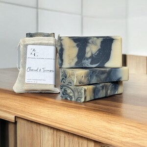 May include: Artisanal skincare soaps with charcoal and turmeric. The soaps have a marbled design with black and tan colors. A small muslin bag with a label that reads "Charcoal & Turmeric" is also in the image. The soaps are 4 oz each.