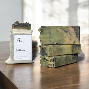 May include: A bar of green, black, and brown soap with a white label that reads "Earthbound" and "100% handcrafted 120 ml | 4 oz". The soap is sitting on a wooden surface.