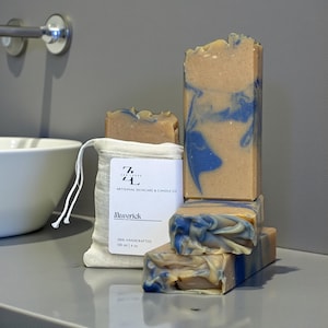 May include: Artisanal skincare soap bars in shades of tan and blue, stacked on a shelf. The bars have a marbled design and are next to a small muslin bag with a label that reads "Maverick" and "120 ml | 4 oz".