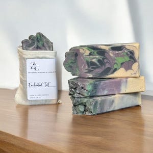 May include: Four bars of handmade soap stacked on top of each other. The soap is a swirl of green, black, and purple. The soap is packaged in a white muslin bag with a label that reads "Enchanted Tart" and "120 ml | 4 oz".