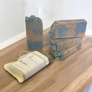 May include: Artisanal soap bars in shades of blue and brown are stacked and displayed on a wooden surface. One bar is packaged in a beige mesh bag with a label that reads "No Lune" and "Artisanal Skincare & Candle Co."