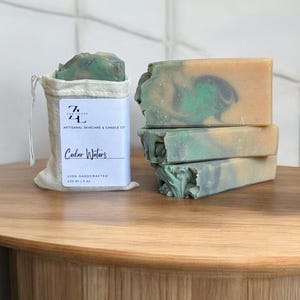 May include: Four bars of handmade soap in a stack. The soap is a light green and beige color with swirls. The soap is packaged in a white muslin bag with a label that reads "Cedar Waters" and "100% Handcrafted 120 ml | 4 oz".