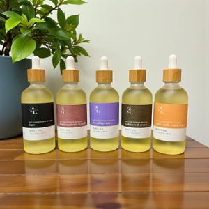 May include: Five glass bottles of body oil with bamboo lids and white droppers. Each bottle has a different label color: black, burgundy, purple, brown, and orange. The labels read "bare", "black raspberry & vanilla", "french lavender", "cashmere & cream", and "goat's milk, oat & honey".