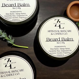 May include: Three round black tins of beard balm with white labels. The labels read "ZEN LUXXE ARTISANAL SKINCARE & CANDLE CO." and "Beard Balm 2oz | 57g." Ingredients include Shea Butter, Mango Butter, and Grapeseed Oil. A sprig of greenery is visible.