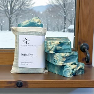 May include: Artisanal skincare soap bars in shades of blue and white, stacked on a wooden shelf. A linen bag with a label reading "Juniper Drift" and "100% Handcrafted" is next to the soap. The soap bars are approximately 4 oz each.