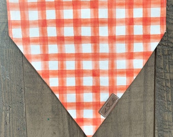 May include: Orange and white plaid dog bandana. The bandana is a triangle shape with a watercolor-style plaid pattern. A small brown leather tag with embroidered initials is attached to the bandana.