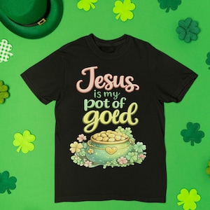 Könnte beinhalten: Schwarzes T-Shirt mit dem Text "Jesus is my pot of gold" in rosa und gelben Buchstaben. Das Shirt zeigt eine Grafik eines Goldtopfes mit einem Herzen und Kleeblättern. Ein grüner Hut und Kleeblattdekorationen sind auf einem grünen Hintergrund verstreut.