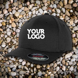 Op de afbeelding: Zwarte Flexfit baseballcap met de tekst "YOUR LOGO" in het wit. De cap heeft een gebogen klep en een Flexfit label. De hoed is maat S/M.