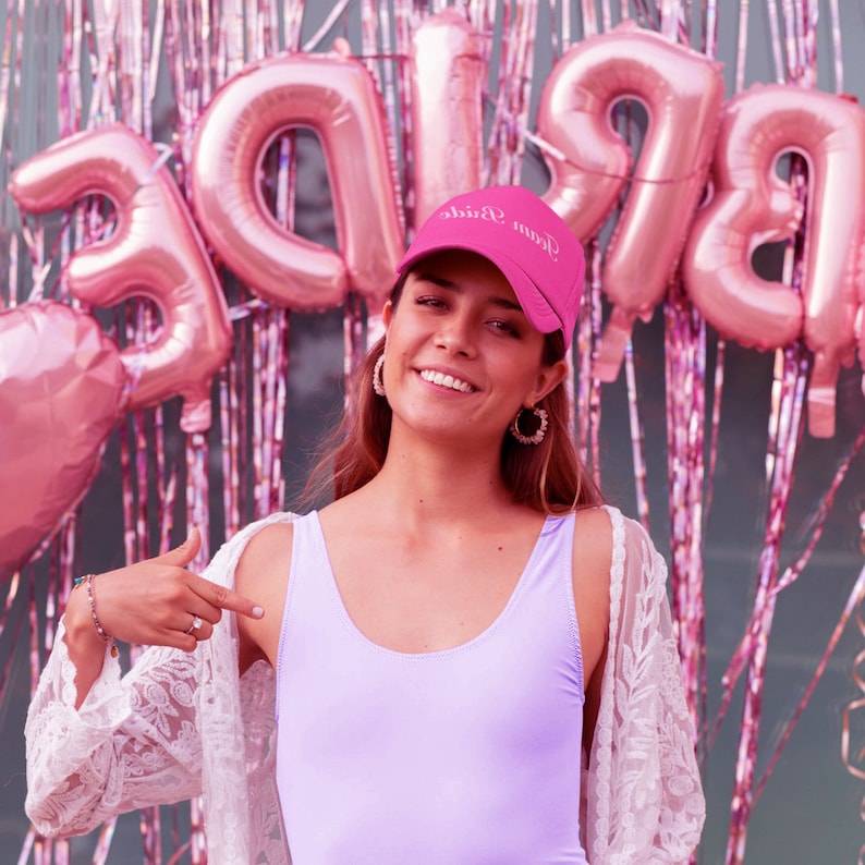May include: A woman wearing a pink baseball cap with the text "Team Bride" in white lettering. She is smiling and pointing to her purple tank top. The background is a wall decorated with pink balloons.