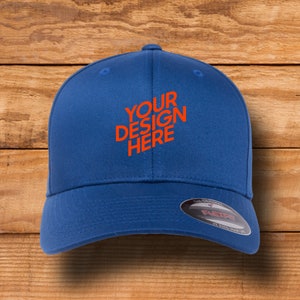 Puede incluir: Una gorra de béisbol azul con visera curva y correa ajustable. La gorra tiene el texto "YOUR DESIGN HERE" bordado en naranja en la parte delantera.