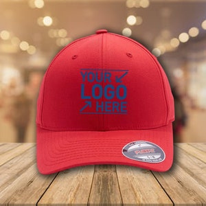 Op de afbeelding: Rode baseballpet met een blauw geborduurd logo met de tekst "YOUR LOGO HERE". De pet heeft een zwart Flexfit-label aan de achterkant.