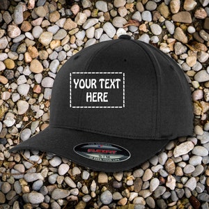 May include: Black baseball cap with a white embroidered box that says "YOUR TEXT HERE" on the front. The cap has a Flexfit label on the back.