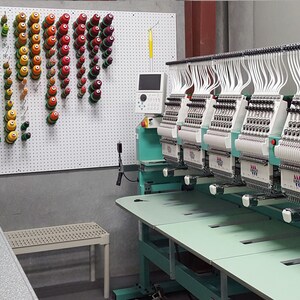 May include: A close-up of a multi-head embroidery machine with a variety of coloured spools of thread hanging on a pegboard in the background.