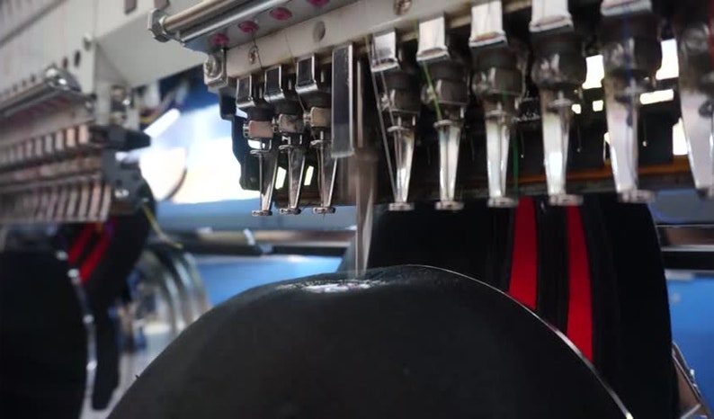 May include: Close-up of a black fabric being embroidered by a multi-needle embroidery machine. The machine has multiple needles and thread spools, and the fabric is being stitched with red thread.