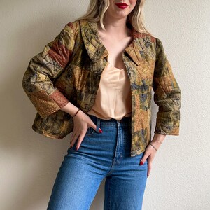 May include: A quilted jacket in a patchwork design featuring shades of olive green, brown, and rust. The jacket has a collared neckline and three-quarter sleeves. The model is wearing blue jeans and a peach-colored top.